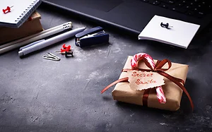 Package marked Secret Santa with brown paper, candy cane and ribbon surrounded by office supplies