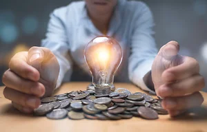Businessman hand protecting heap of coins and lightbulb which glowing. Creative new business idea can make profit money concept.
