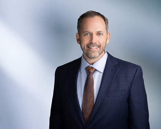 Man, Mikhail Murshak, with short dark hair wearing a navy blue blazer, smiling slightly against a blurred white and blue background.